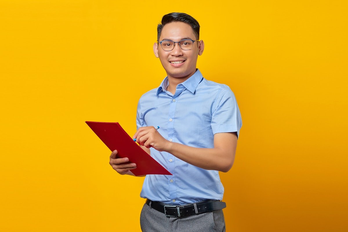 Homme asiatique souriant, portant des lunettes et une chemise bleue, tenant un dossier rouge.
