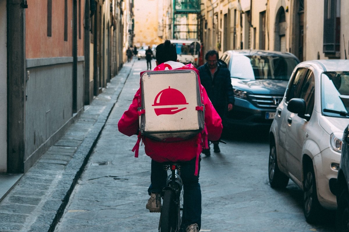 Livreur à vélo avec un sac à dos rouge dans une ruelle urbaine.