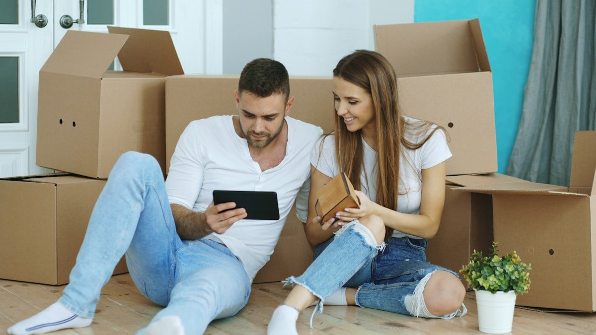 Jeune couple assis sur le sol entouré de boîtes, utilisant des appareils électroniques.