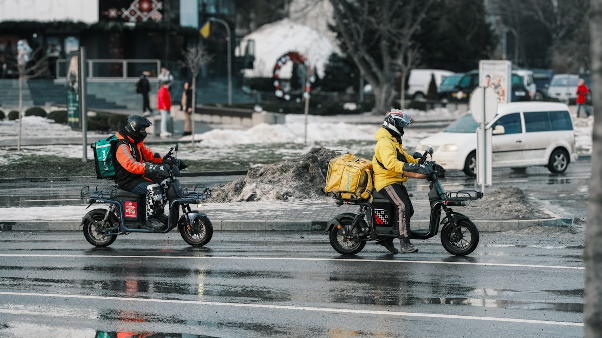 Deux livreurs à scooter électrique traversent une rue enneigée.