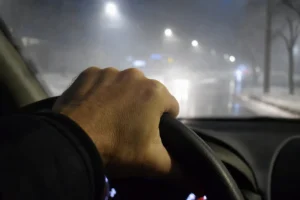 Main serrée sur volant d’une voiture, rue enneigée, lumière des phares, sécurité routière en conditions glacées.