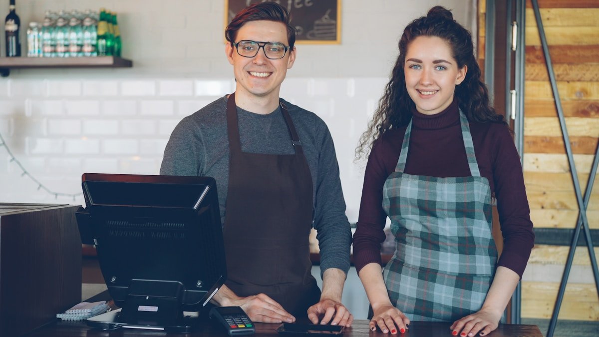 Deux employés souriants dans un café, l'un portant un tablier à carreaux.